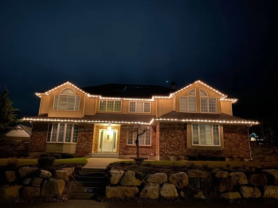 Two-story stucco and brick home outlined in warm white C9 lights above a rock wall